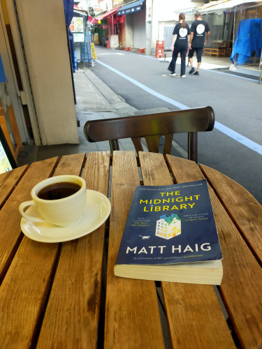 Photo is of the outside of a cafe looking on to the alley which is bordered by thick blue lines. Two people in black can be seen from behind walking in the distance. In the foreground is a faux wood-slatted (perpendicularly) table with a white mug & saucer of black coffee to the left and the dark blue paperback book to the right with a paper folded building illustration in the center, with a white shadow cat to the left of it