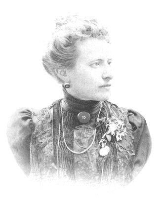 Blanca Catalán de Ocón y Gayolá in black and white head and shoulders photo. Late C19th /early C20th high necked dark blouse with lace on front and a round brooch at neck. hair swept back and pilled high at back, half profile, looking to picture right