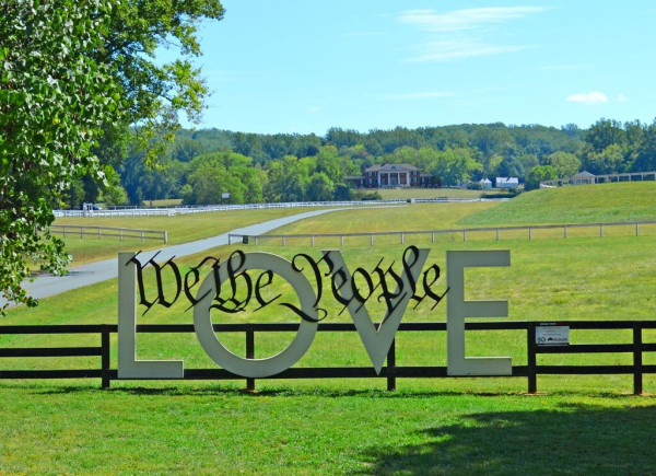 A Virginia LOVEWORKS sign with the words "We the People" placed over the top of the word LOVE. The Madisons' mansion and the white South Yard buildings can be seen in the background. The grass and trees are a bright green, the sky a light blue with few clouds. 