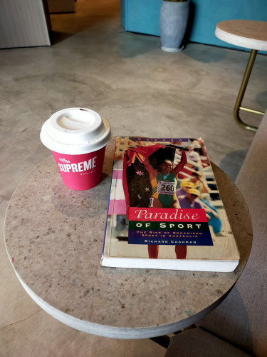 Photo of inside of cafe. On the round Formica table is a red paper coffee cup with a white lid. On the cup in white reads Coffee Supreme. To the left is the paperback book which features a photo of Aboriginal Australian Olympic Gold Medalist Cathy Freeman in a green and yellow bodysuit holding the Australian flag below the Aboriginal flag