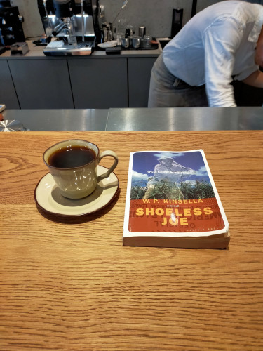 Photo is of a wooden cafe counter. The paperback book is on the counter. It features a red bottom section for the author/title mentioned in the skeet. The top part is a ghostly baseball batter in a cornfield with a blue sky. To the left is a greyish tan coffee mug and saucer of black coffee. Behind is the headless barista in a dress shirt bending over. Behind him are various coffee shop implements including a small espresso machine