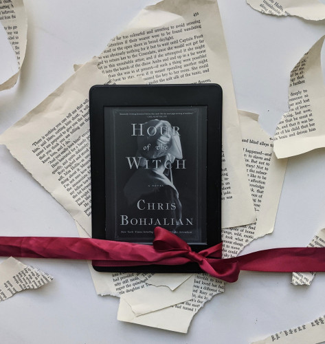 My Kindle, showing the cover of The Hour of the Witch by Chris Bohjalian. A maroon/scarlet ribbon is tied across the bottom and several ripped up book pages are in the background.