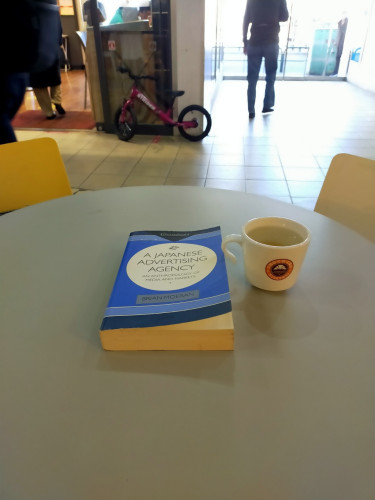 The photo is of a blue paperback book with white circle for the title on a round grey outside cafe table. To its right is a white mug of yuzu tea. The mug has the orange ring around a brown circle in which is a white croissant illustration that is St Marc's Cafe's logo. In the distance you can see the legs of pedestrians a a small red bike for a child up against a wall
