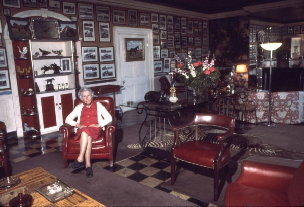 Photo courtesy of The Montpelier Foundation. An elderly woman sits with her legs politely crossed, her hands folded on her lap. She wears a red dress, black shoes, and white cardigan. She sits in a lavishly furnished room, well decorated with photos of horses filling the wall. A cabinet of statues and trophies stands behind her. There is a brightly colored bouquet of flowers in the center of a glass table, a zebra skin rug, black and white checked floors, and red accents throughout the room.  