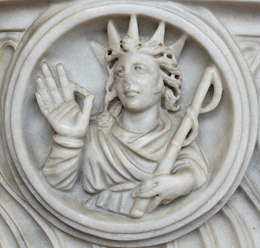 Marble relief of Sol-Helios. The sun god is crowned with a five-spiked sun ray crown and he is wearing a cloak pinned over his right shoulder. He is holding up his right hand with the palm towards the viewer, cradling a whip in his left.