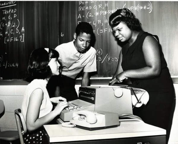 Black and white image of a student at an IBM terminal with another student looking on as a teacher explains how to use the machine. A blackboard with mathematical notation is on the black board.