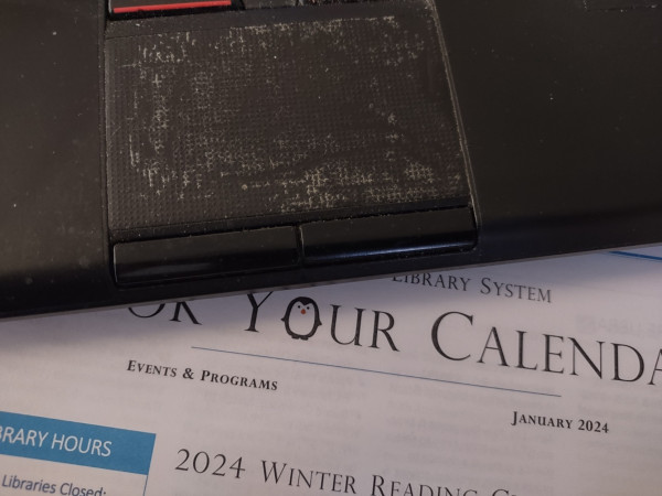 Photo: Part of the keyboard, including the touchpad of a well-used Lenovo laptop with a printed sheet of paper sticking out from underneath. Some of the words on the paper are visible:
"Library System
Book Your Calendar
Library Hours
2024 Winter Reading Challenge"