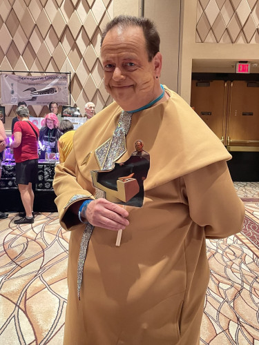 Picture of Blake Brocksmith, the son of actor Roy Brocksmith, cosplaying as his father's guest Trek character, Sirna Kolrami.  Dressed in yellow costume with ornament in the front, holding a picture of a Trek captain's chair.