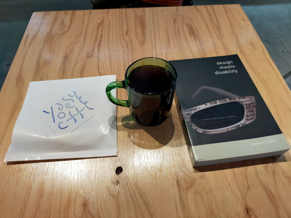 Photo is of a square wooden table inside the cafe. On the left is a square paper with YAESU COFFEE logo of blue text where the O & final E are in between the words YAESU CFFE. In the middle is a plastic green mug of black coffee. To the right is the black paperback book with a single lens of a grey framed sunglass, the frames with black text on it.