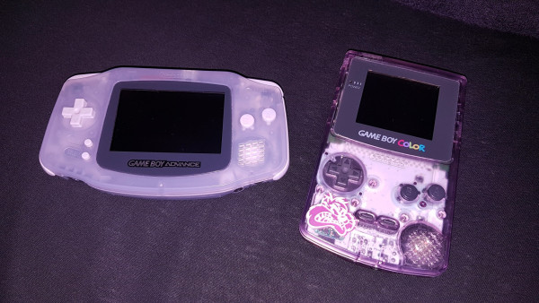 A purple Game Boy Advance on the left and a purple transparent Game Boy Color on the right. The right GB has a purple "glow in the dark" sticker on it of the WB Tazmenian Devil, Taz.