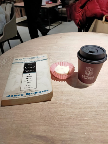 The photo is of a round wooden table. On it is the white paperback book. To the right is a pink cupcake paper in which is a white cheesecake shaped like a cat's head. Further to the right is a brown black-lidded coffee cup with the YANAKA COFFEE logo on it.