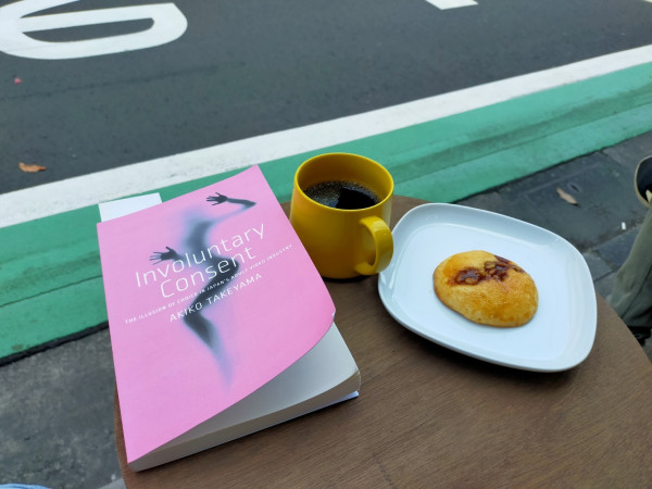 Photo is outside the cafe where a street can be seen with a green stripe against a white stripe. On a brown wooden round table is the pink paperback book with a woman's black naked silhouette. To the right is a mustard yellow mug of black coffee. Further to the right is a square white plate with a tan cookie with a strip of pecans in the middle.