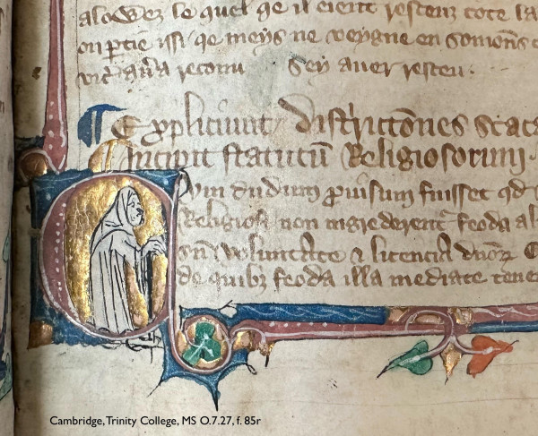 Detail of the lower left corner of a leaf in a medieval manuscript: folio 85 recto in Cambridge, Trinity College, manuscript O.7.27. 3 lines of French, followed by 2 larger heading lines of Latin, then 4 more lines of Latin, all copied in brown ink. The bottom-most 4 lines are indented to accommodate an enlarged decorative initial ‘C’ painted in pink atop a square blue background. Bars of pink, blue, green, and gold extend from the background to form a border around the text. Green and orange leaves spring from the bars. The initial ‘C’ is almost entirely infilled with a background of smoothly burnished, shining gold leaf. The non-gilded negative space forms the shape of a standing monk or friar, depicted in profile, his features and the draping of his robes drawn in with black ink. The tiny fellow gestures towards the adjacent text, gazing upon it morosely. 