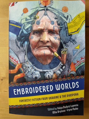 Front cover of a paperback book. Title is Embroidered Worlds. subtitle Fantastic fiction from Ukraine & the diaspora. Edited by Valya Dudycz Lupescu, Olha Brylova, Iryna Pasko. Cover image is a watercolour style of a older person with a strange mechanical contraption around their face, arcane techno tattoos, and an old wooden and gold pipe in their mouth.