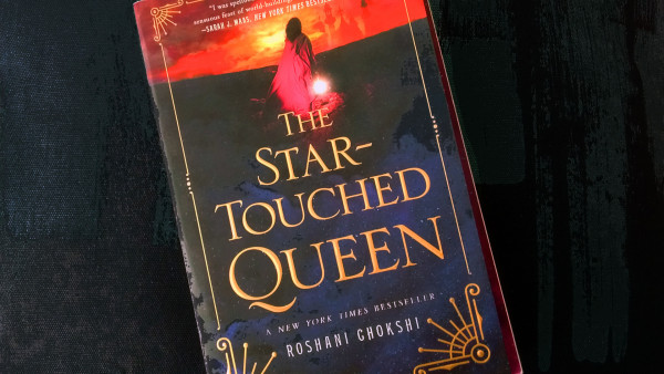 The cover of The Star-Touched Queen by Roshani Chokshi shows a young woman -she wears a Sari, we only see her back- staring into the horizon. It's near sunset, the sky is covered in clouds reflecting the light of a dying sun into a myriad of oranges. The young woman carries a lamp, which light shines the brightest. She stands on dark ground. In gold letter, the title of the book covers that ground.