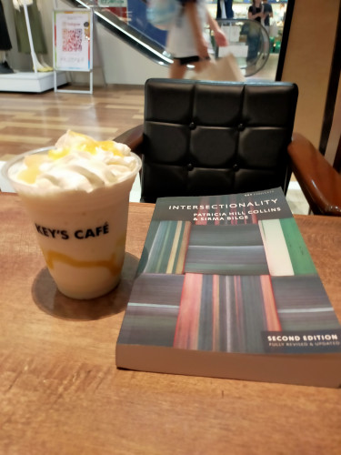 Photo is of a brown cafe table with an upholstered black chair back with thick wooden arms. On the table is the paperback book, black with squares of various color stripes, alternating between vertical & horizontal. To the left is a plastic cup with KEY'S COFFEE on it. Inside is a white shake with yellow line of pineapple. There is whipped cream on top with pineapple pieces  In the distance is the midsection of a woman in black shorts and a white t- shirt walking in front of an escalator