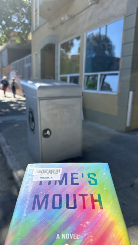 Cover of Edun Lepucki’s Time’s Mouth next to a grey metal book return bin on the sidewalk in front of a storefront with large windows of a beige building.  