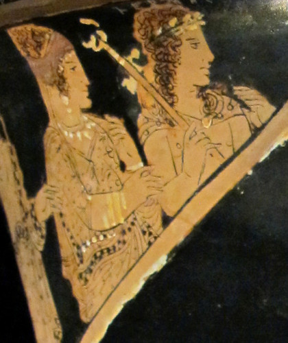 Red-figure vase painting of Hermes depicted with shoulder-length curly hair adorned with a laurel wreath. He wears a cloak around his neck and holds his kerykeion staff in his right hand. Beside him, a goddess with beautiful jewellery is watching the scene as well.