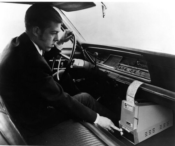 Black and White square image. A man sits in his car and operates a small mobile fax machine that rests on the floor below the dashboard, ca. 1971.