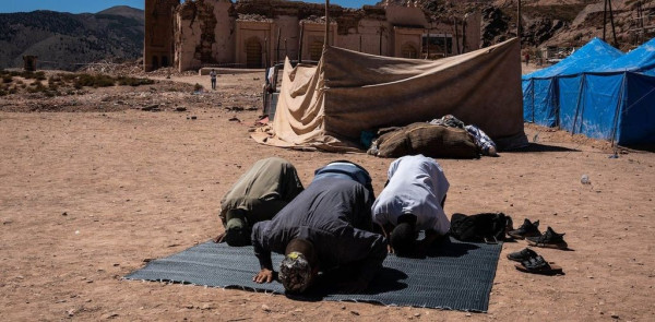 Three people prostrating in prayer on a mat. In the backdrop are mountains and a damaged ancient brick structure.