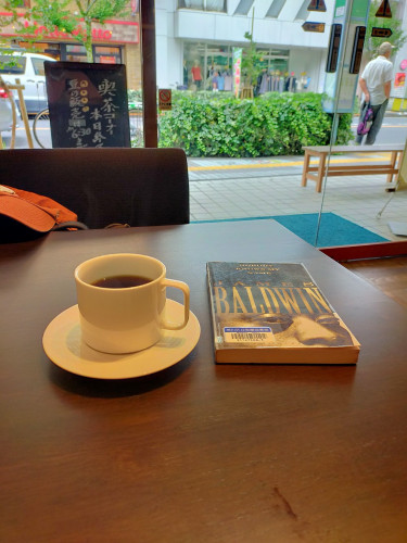 The photo is on a table inside the cafe. There is a white mug and saucer of black coffee. To the right is a black paperback library book with Black author James Baldwin's face looking at the viewer. In the distance outside the cafe is a sidewalk with visible green shrubbery. To the left an old Japanese man is standing by a bus stop