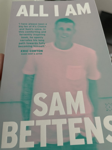 Een foto van het boek All I Am van Sam Bettens. De cover heeft de titel aan de bovenkant van het boek en de naam van Sam aan de onderkant, beiden gecentreerd in witte tekst. De cover is een foto van Sam in groen/roze pastelkleuren. De foto is ietwat vaag.