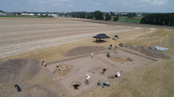 what looks like a fallow field with a large rectangular archaeological dig at center bottom. there are several people standing and digging around the site.