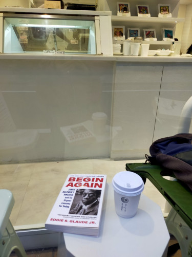 The photo is of seating outside a cafe against a large window. At the top you can see the ice cream area and shelving of coffee bean bags above paper cups & other cafe produced detritus. There is so much white. Outside on a tiny round table is a white coffee cup with a white lid. The white paperback book mentioned in the post has BEGIN AGAIN & the author's name in red. There is a black& white photo of the Black writer James Baldwin looking at the camera
