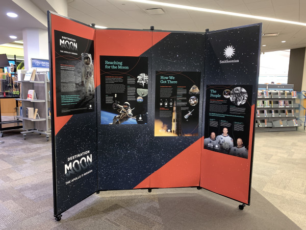 Temporary exhibition wall set up in the middle of a library. The panels are 5 feet high and are covered completely with  museum-style exhibition of text and images of American astronauts. A logo that reads Destination Moon is visible in one corner. The Smithsonian logo is visible in the opposite corner.