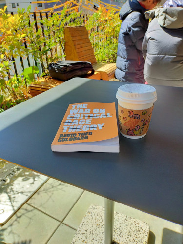 The photo is of a square silver metal table of ant outside seating area. The orange paperback book mentioned in the skeet is on the table. The title is in white bold letters with a black line through CRITICAL RACE THEORY. The author's name is below the title in smaller bold black letters. To the right is a white paper lid & cup of coffee with a brown sleeve with little designs on it of birds & other shapes. In the distance you can see a black computer bag on a bench and 2 other Japanese patrons in (left to right) a puffy black winter jacket and puffy grey winter jacket.
