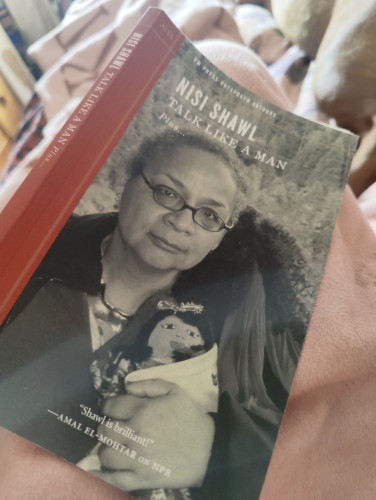 Book laying over my lap opened with the cover showing. Cover has a Black woman with a doll in her hand. Title is Talk Like A Man by Nisi Shawl.