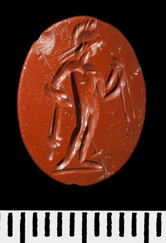 Oval intaglio of blood red jasper with the figure of Mars skilfully etched into it. He is standing in the nude, only a crested helmet on his head the folds of a cloak over his right arm in which he cradles a long spear. In his left he holds a sheathed sword with its belt. His head is turned to his left, looking at the sword.