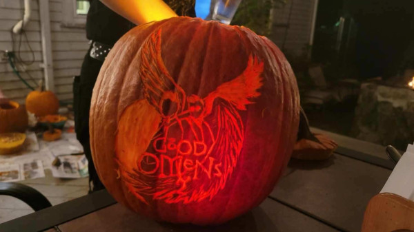 A pumpkin carved with a partial-thickness engraving of an interpretation of the Good Omens Season Two poster. There is a person's arm stuck into the top of the pumpkin. There are several partially-disemboweled pumpkins visible in the left-hand background, and a fire pit on a stone well is just visible to the right.