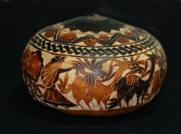 A gourd that had been decorated with carvings of two llamas or alpakas, some plants in the background, and a human standing left of the animals.