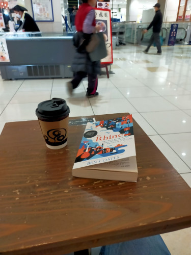 The photo is of a wooden table on which is the paperback book with a white space representing The Rhine river between colorful buildings & mountains representing the major cities along the river. To its left is a paper black plastic lidded brown sleeved coffee cup. A few blurry Japanese people can be seen in the distance of a mall