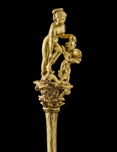 Photo of a long and elegant gold hairpin, adorned with a charming figurative scene: positioned at the top of a Corinthian capital featuring an abundance of acanthus leaves, a small Eros offers the necessary support to the goddess Aphrodite who rests above him, her foot balanced over a dolphin. Eros holds a mirror in his hands as the nude goddess appears to adorn herself, adjusting one of her anklets to complement the snake-shaped arm bracelets she already wears. With further fine incisions decorating the entire length of the pin, this luxury object can be clearly linked to its use as stylish ornamentation.