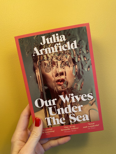 a photo of the cover of Our Wives Under The Sea by Julia Armfield. on the cover there's a young white woman facing us, on a green-grey background, we can only see her face and naked shoulders. her features are distorted as if there's a glass covered with wet, melting ice between us and her. she appears to me somewhat lost or longing for something, as if she is confined in this icy glass box