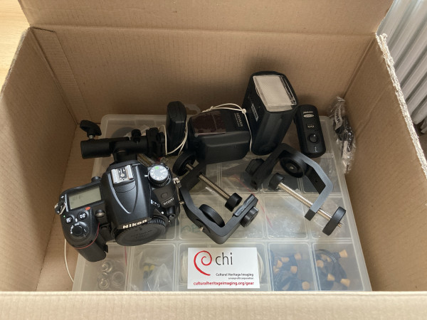 A cardboard box filled with various photography equipment including a camera, flash units, and accessories.