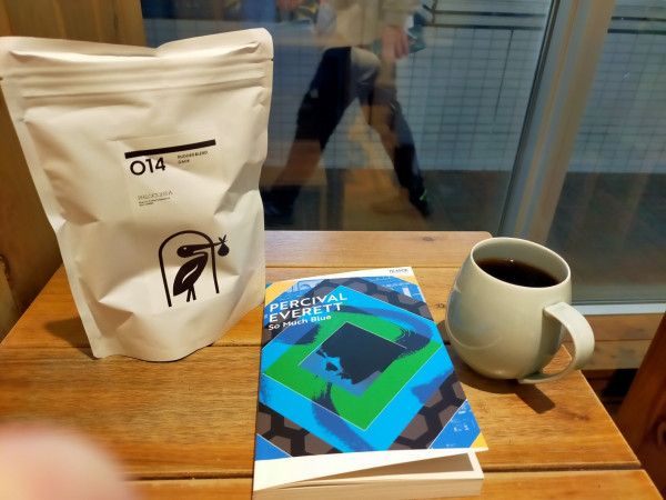 Photo is of a wooden table. In the middle is the blue paperback book. To the right is a gray mig of black coffee. To the left is a white bag of coffee beans featuring the pelican logo holding a bag in their beak. Across the way is a window, where we can see a blurry Japanese man walking past to towards the left