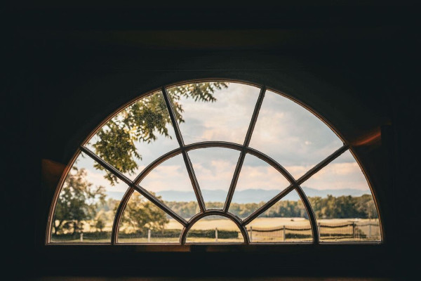 A half-circle window looks out onto blurry mountains, a thick tree line, grassy field, and fence. 