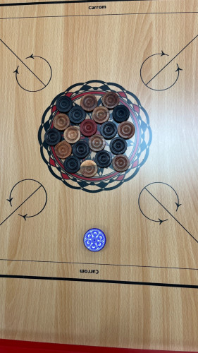 Picture of a Karrom board. Tan coloured surface with a black square within another black square. Above and beneath are typed the words "Carrom". In the middle is a patterned circle with a red Queen disc and 18 discs (9 black, 9 tan) arranged in alternating succession. Directly below is the lilac "striker" disc. Jutting from each angle is a line closed by a semi circle, meant to indicate points where shots can be made. The object of the game is to use the striker to get your disc (depending on colour) onto one of the four pockets. If you get one in, you get another shot. If you hit the queen disc into the pocket, the next shot has to be your own disc. If you do this, you win. If you don't, the queen returns to the centre of the board.