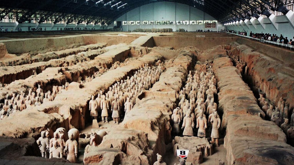 Scene of the Terracotta Warriors in China in rows in the ground in situ. The Dig is surrounded by a ring of tourists within the large shelter. 