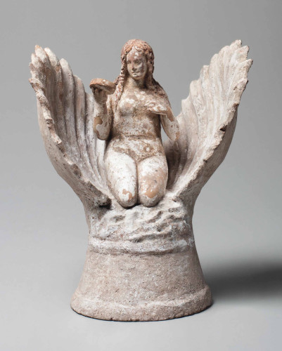Photo of a terracotta figurine depicting the birth of Aphrodite. The goddess emerges from an open scallop shell, depicted nude, seated with her legs tucked beneath, her long wavy hair falling onto her shoulders, looking at her reflection in a mirror held in her right hand, on a high cylindrical plinth. Some polychromy and white slip are preserved throughout. The nude goddess is raising her right hand holding her mantle, a dolphin below to her right, her lowered left hand on a column, preserving some pink and red pigment throughout.
