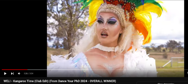 Still from linked video, shows a drag queen looking surprised in a flamboyant feathered costume. 
