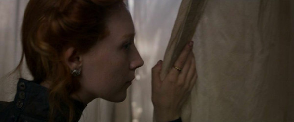 A redheaded woman pulling back a pale curtain