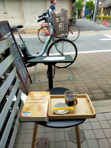 Photo is of outside a cafe. The large orange hardcover library book is on the small round black metal table . To the right is a wooden tray on which is a clear cup of black coffee with a brown plastic handle. In front is a white plate with 2 yellow sugar sticks perpendicular. To the left is a green slated fence. In the distance is a parked turquoise electric bicycle with a grey collapsible stool in the front black basket. In the further distance is a Japanese woman pedaling to the right on her bicycle