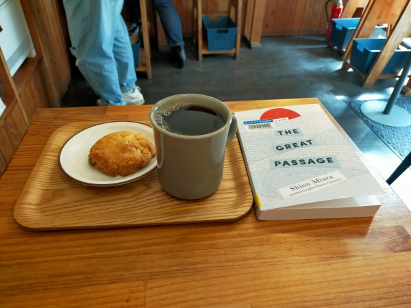 The photo is of inside cafe. On a brown wooden square table is the paperback library book which is white with segments of text looking like waves, above which is a half red circle resembling the flag of Japan and a sun. To the right is a brown wooden tray on which is a grey mug of black coffee. To the left on a small round white dish is a tan cookie. In the distance we can see the turquoise jeans of a patron and regular dark blue jeans of another. We can also 3 blue buckets for patrons bags underneath wooden stools.