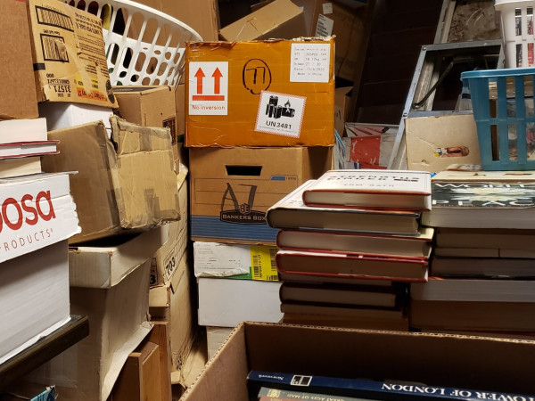 A photo of a cramped storage area with dozens of mismatched cardboard boxes, seeming haphazard in their arrangement – to the uninitiated. Within some of the boxes and stacked in open spaces are books of all sorts and sizes. Other boxes contain movies, comics, and magazines. Many of the boxes have been emptied and piled out of the way for future re-use. Others hold work to be done, prior to display, plus a few baskets of items ready to go. An ancient aluminum folding ladder leans against a temporarily unused magazine display rack in the background.