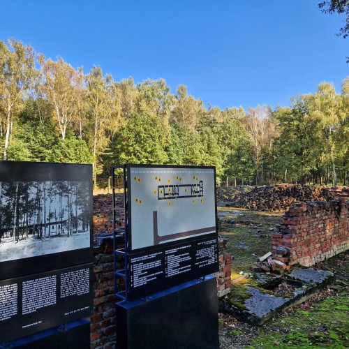Ruis of a blown up gas chamber and crematoria. Bricks forming a structure located between trees. In the foreground - information plaques about the building.