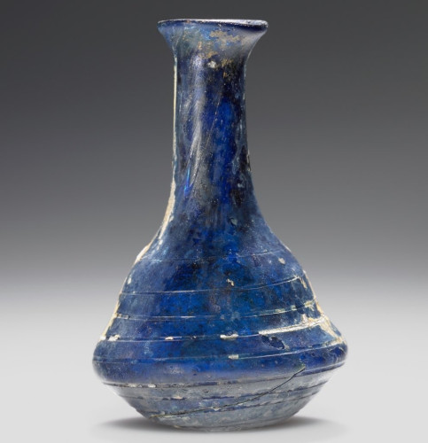 Description from museum: “An ointment flask of free-blown opaque, dark-blue glass. It is decorated with grooves encircling the body. The lip is slightly lopsided. Some weathering has given the vessel a blue-purple iridescence. There is also some incrustation.”